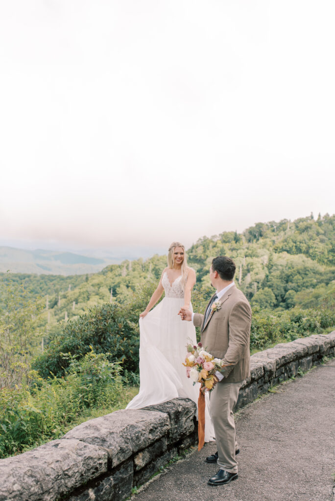 Bride walking along a stone wall overlooking a scenic mountain vista in the Blue Ridge Mountains holding her grooms hand. Groom in a tweed suit holding autumn colored bouquet