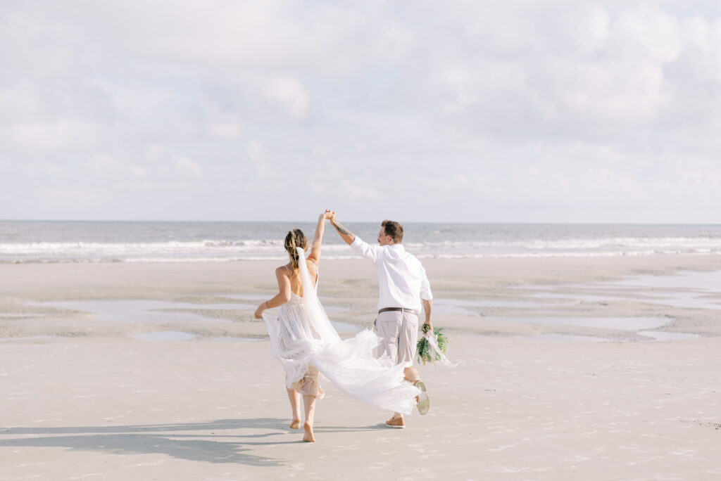 Bride and groom running joyfully towards the ocean on the beach after their elopement, bride in an ivory lace dress and groom in shorts and a white shirt on Fripp Island in South Carolina