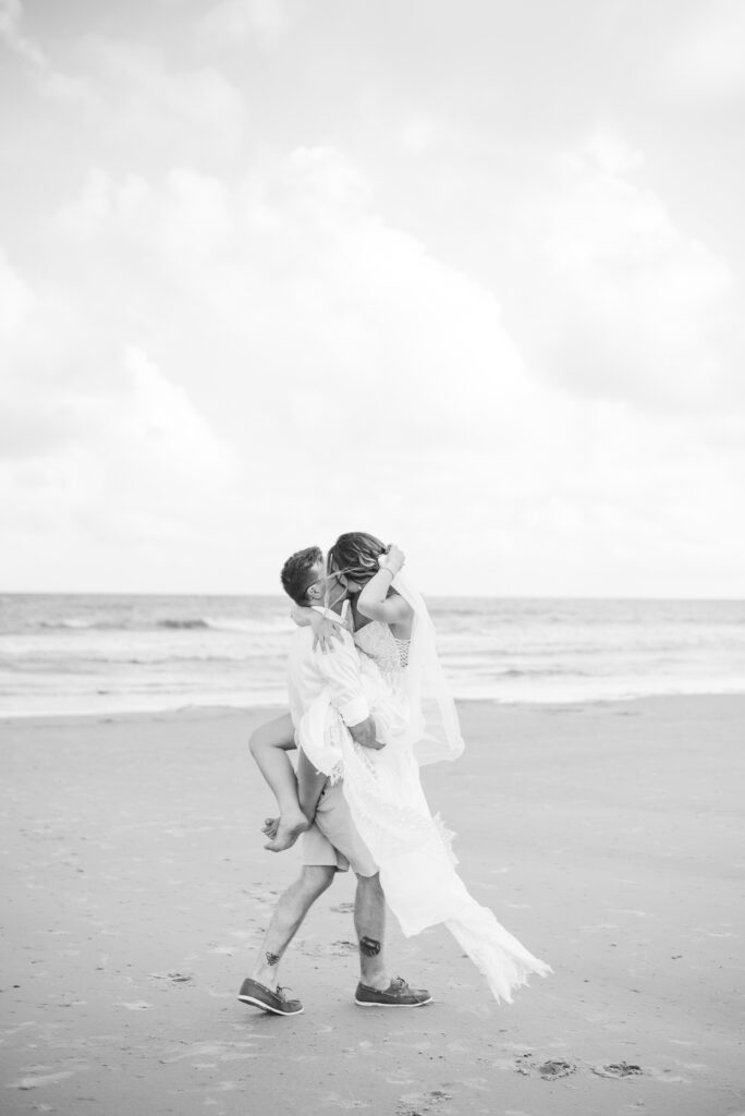 Groom holding his bride with her legs around his waist kissing on the beach in black and white