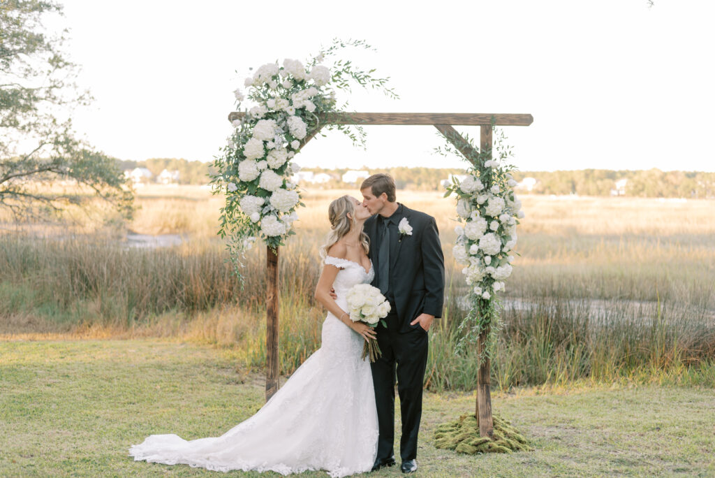 Bride and groom kissing under their ceremony arch at sunset in front of the marsh at a private home wedding in Charleston South Carolina