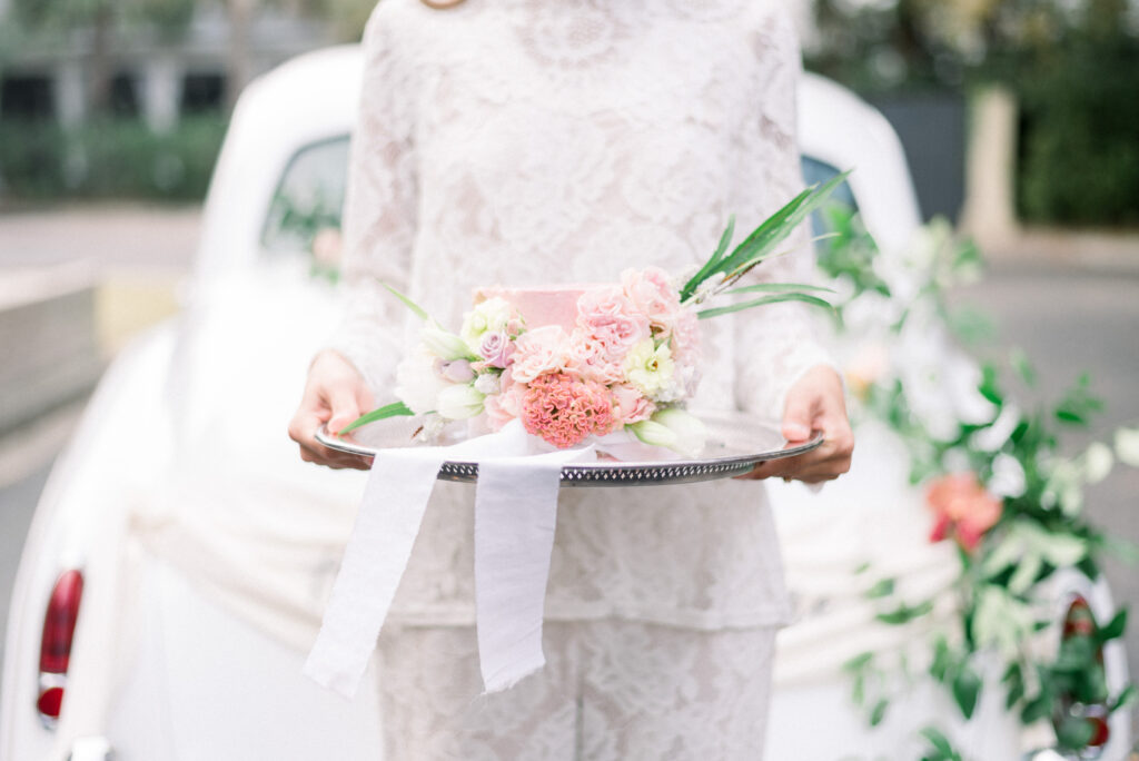 Bride in a lace pants suit holding a pink floral mini cake during micro wedding in Charleston