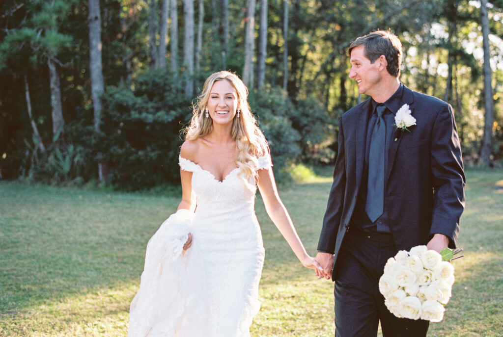 Bride and groom walking holding hands with white long stem rose bouquet at sunset