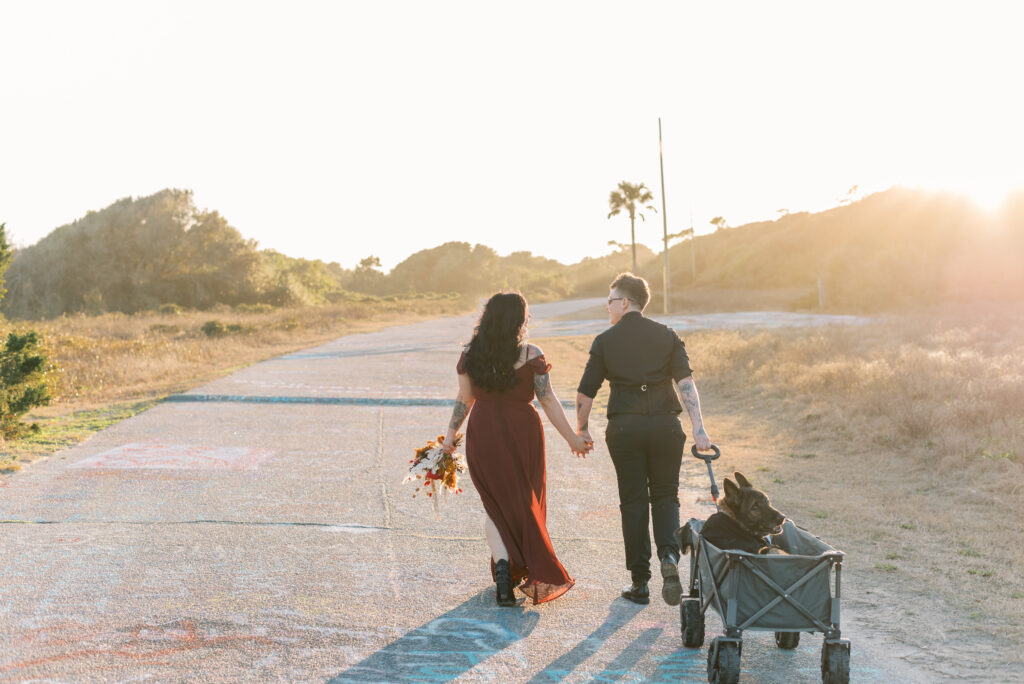 Bride and bride wearing black and burgundy pulling a dog in a wagon walking holding hands into the sunset along a graffiti path