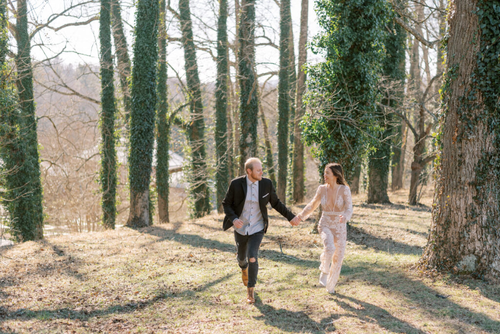 A bride and groom are captured in a photograph, holding hands and joyfully running through the woods at the Biltmore estate in Asheville, North Carolina. The bride is dressed in a stunning ivory lace jumpsuit, with her hair flowing down in loose waves. The groom is dressed in a black jacket and boots, paired with a light-colored shirt. Their outfits blend seamlessly with the natural setting, as they dash through the trees and over leaves on the forest floor. The image captures the joy and playfulness of the couple, as they celebrate their special day in a picturesque and serene location.