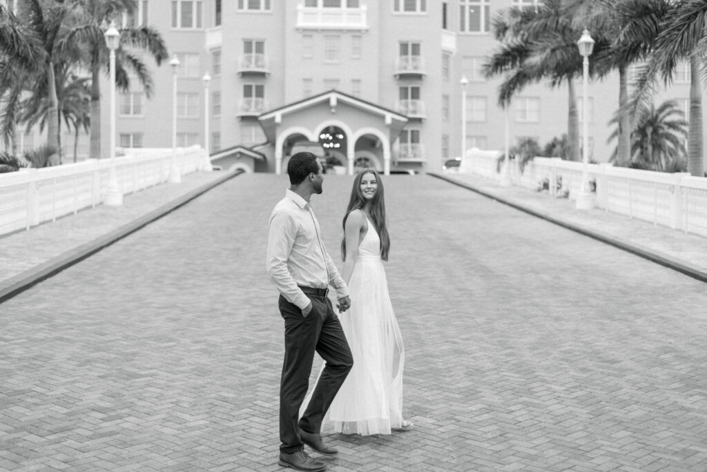 A man and woman are captured in a black and white photograph, walking hand in hand in front of the iconic Don Cesar hotel in St. Petersburg, Florida. The woman is wearing a flowing white dress, while the man is dressed in a dark pants and light shirt. They both exude an air of sophistication and grace as they stroll along the sidewalk, enjoying each other's company. The grandeur of the hotel's architecture looms in the background, adding to the timeless elegance of the scene.