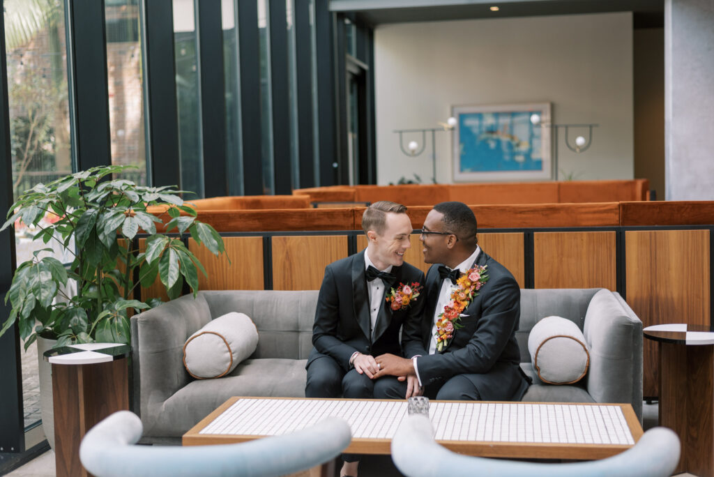 Two grooms are seated side by side on a modern couch at Hotel Haya in Tampa, Florida, on their wedding day. They are holding hands and smiling happily, dressed in elegant black tuxedos adorned with stunning floral pieces. The grooms radiate joy and love as they celebrate their special day together. The stylish backdrop of the hotel's interior adds a touch of sophistication to the image, as the couple basks in the warmth and love of their union.