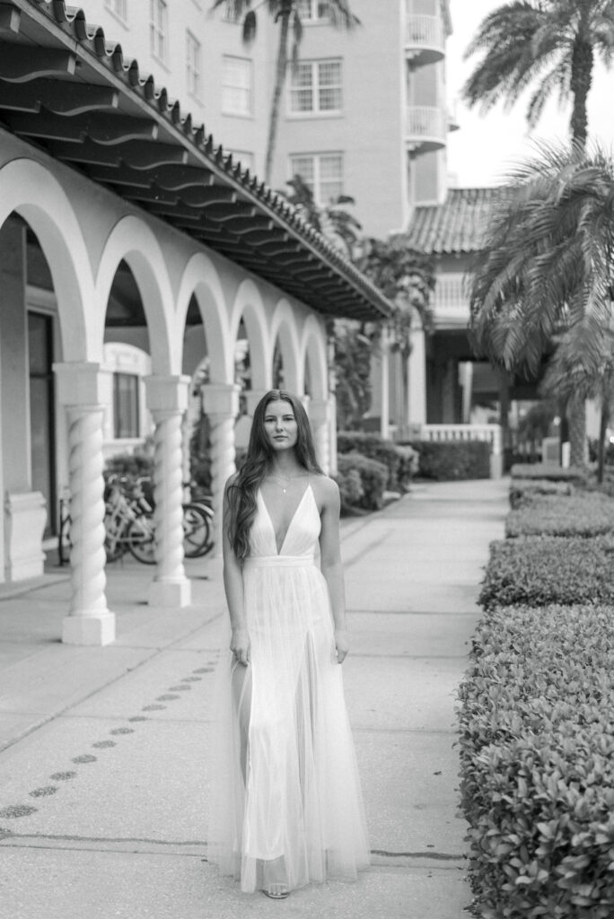 A woman is depicted in a black and white photograph, standing confidently in front of the Don Cesar hotel in St. Petersburg, Florida. She is wearing a sleek and stylish white spaghetti strap dress with a deep V-neckline that accentuates her figure. The dress flows elegantly around her legs, and her hair is down in loose waves. The woman's stance exudes confidence and poise as she gazes off into the distance. The grandeur of the hotel's architecture provides a dramatic and timeless backdrop to the image.