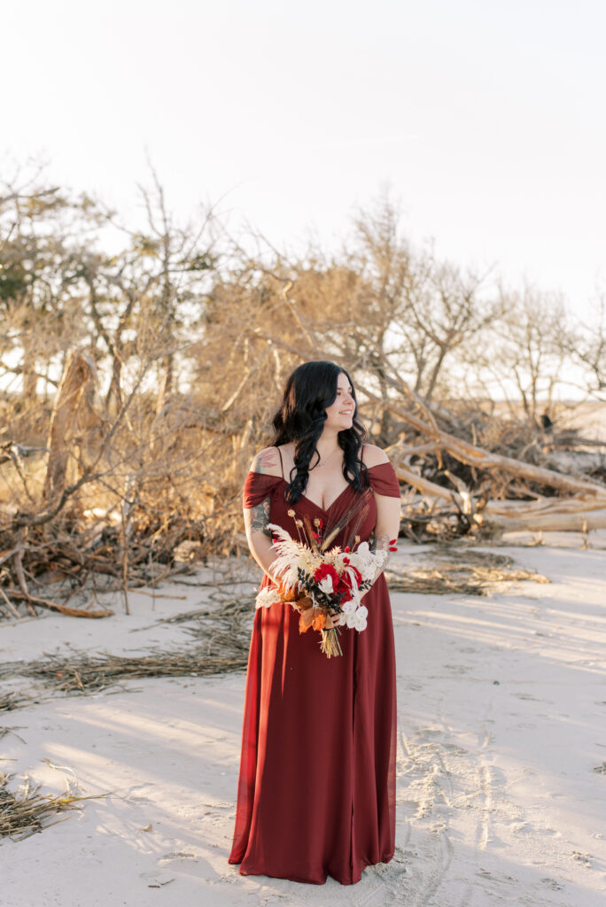 A bride stands alone on a sandy beach, holding her elopement bouquet in both hands. She is dressed in a beautiful burgundy gown that flows gracefully around her feet. The gown features a flattering off-the-shoulder neckline. The bride's hair is styled in loose waves that catch the sea breeze. The soft colors of the sand, ocean, and sky create a tranquil and romantic atmosphere around her. This image captures the quiet beauty and intimacy of an elopement on the beach.