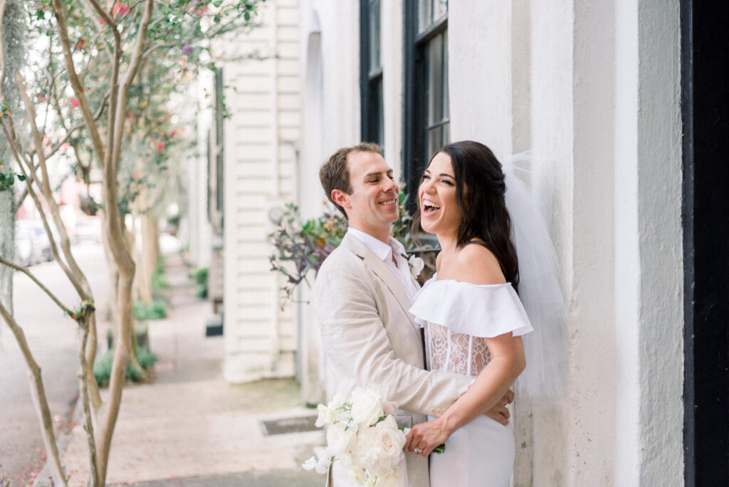 A joyful bride and groom share a moment in historic downtown Charleston, standing on a sidewalk against a beautiful historic building. The bride is wearing a stunning tea-length corset dress and laughing, while the groom smiles at her in his khaki suit. Pink crepe myrtles are in full bloom adding to the romantic atmosphere. Photographed by Kelsey Halm The Wild Elopement