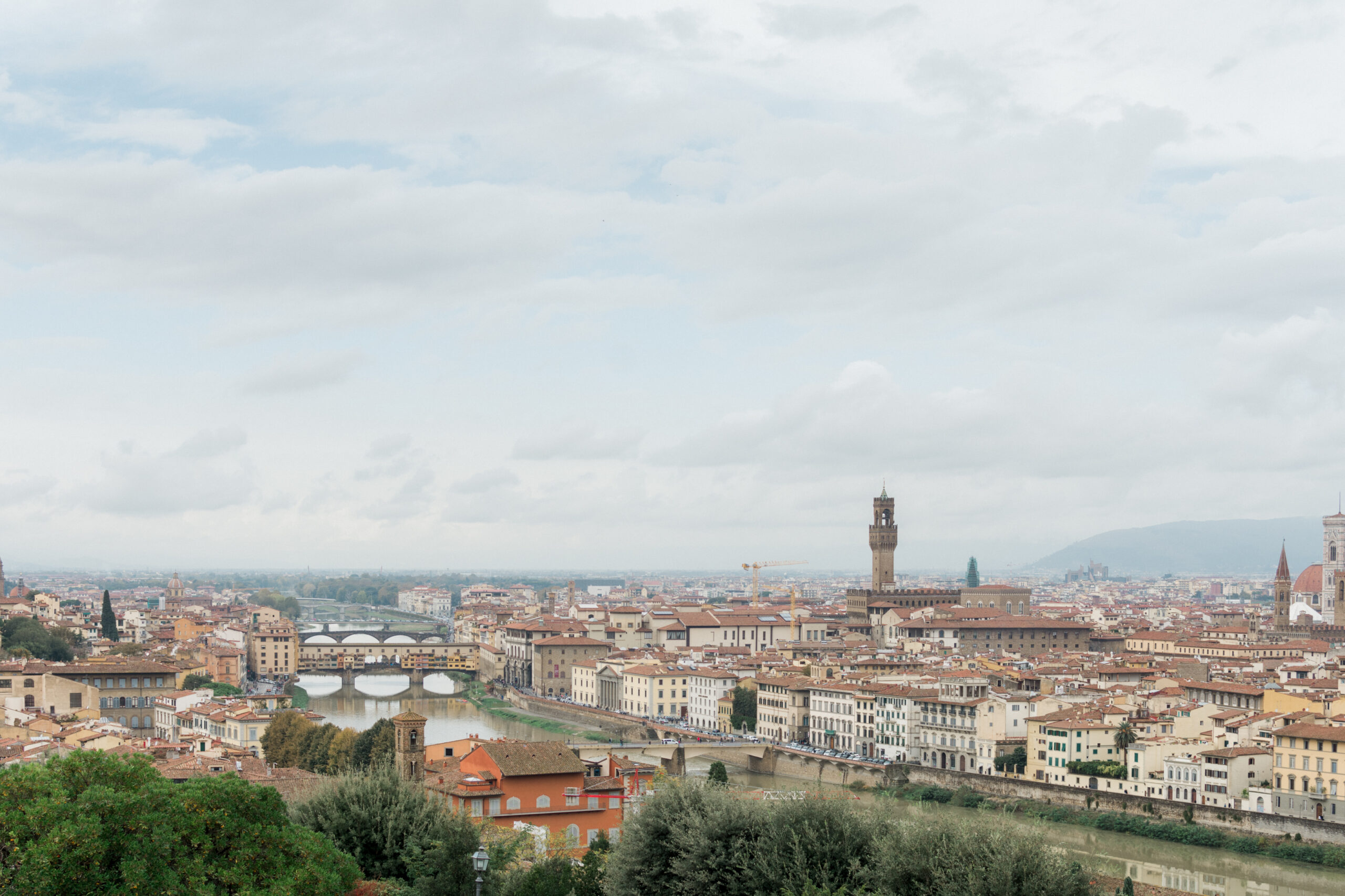 A panoramic view of Florence, Italy with the Arno River in the foreground and two bridges in the distance. The cityscape features historic buildings with red roofs and the famous Duomo cathedral in the center.