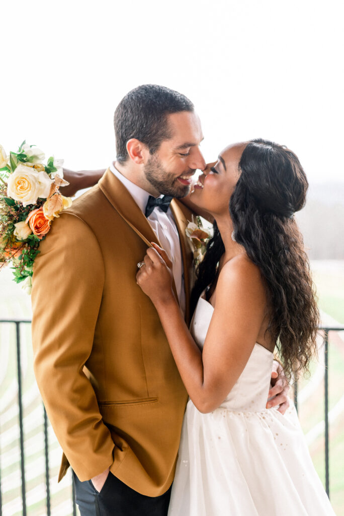 Joyful biracial couple on the verge of a kiss: This heartwarming image captures a radiant biracial couple sharing a blissful moment. The groom looks dashing in his mustard-colored dinner jacket paired with black tuxedo pants and a classic black bowtie. The bride exudes elegance in her strapless white gown, holding a vibrant bouquet adorned with a delightful mix of orange, green, yellow, and peach flowers. The anticipation of their kiss is palpable, reflecting the love and happiness they share. This enchanting photograph beautifully captures the couple's unique style, their affectionate connection, and the vibrant colors that add a touch of vibrancy to their special day.