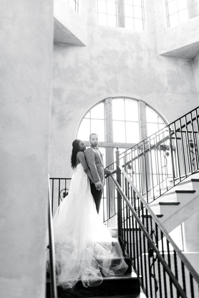 A romantic black and white image of a bride and groom standing together on the grand stairwell at Montaluce Winery in Georgia. The bride is wearing a beautiful strapless gown with a long chiffon skirt that drapes on the stairs, while the groom is dressed in black tux pants and an elegant mustard tux jacket. The epic stairway has iron railings and stucco walls, creating a dramatic and timeless setting for this intimate moment between the couple.