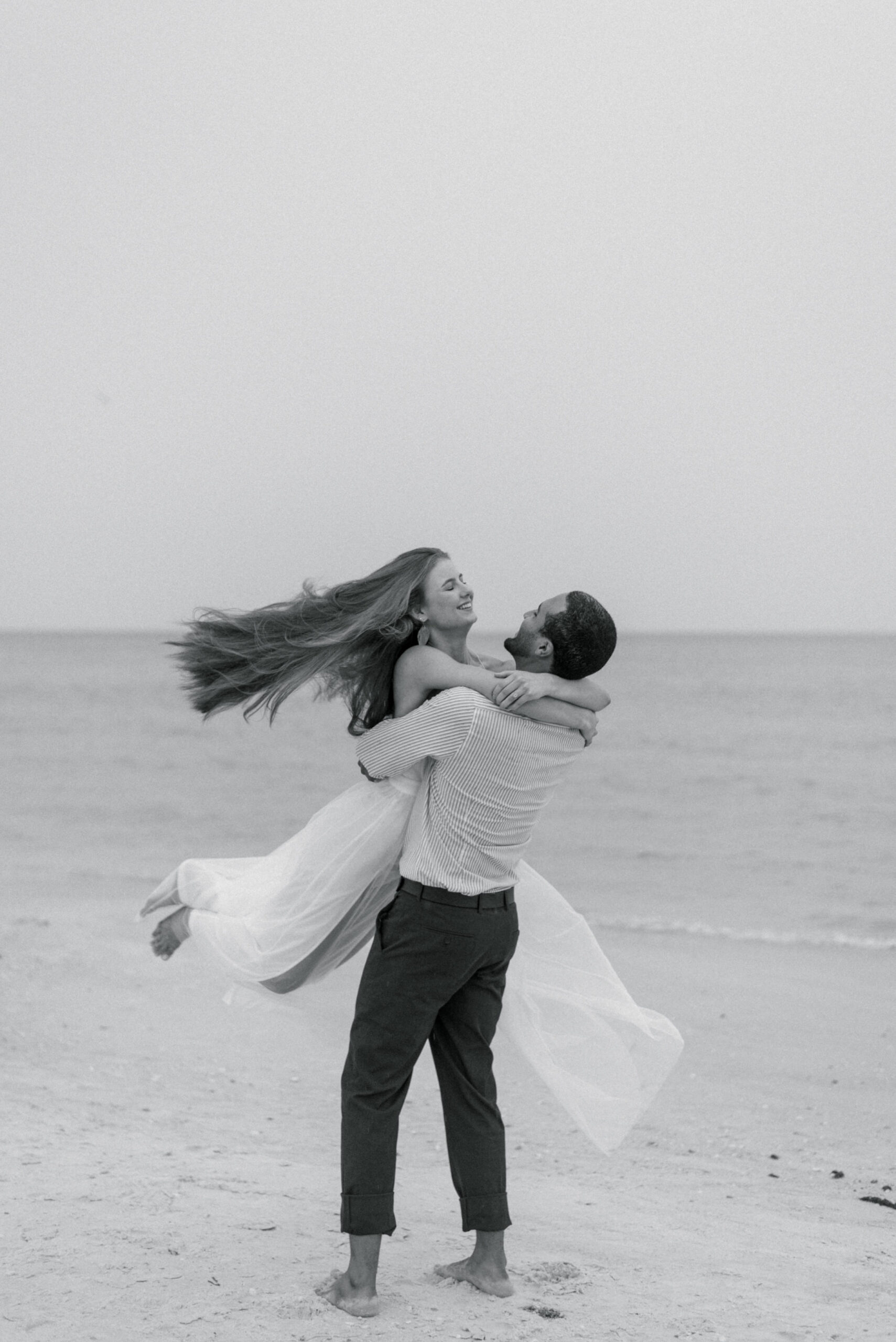 black and white photograph, the groom joyfully lifts and spins his radiant bride on the sandy shores of Saint Petersburg Beach, Florida. The picturesque scene unfolds against the backdrop of glistening waters, creating a timeless moment of pure bliss. The bride, dressed in a ethereal white spaghetti strap dress, exudes elegance and grace, while the groom complements her perfectly in his smart slacks and button-up shirt. This enchanting beachside celebration captures the essence of their love and the breathtaking beauty of their surroundings.