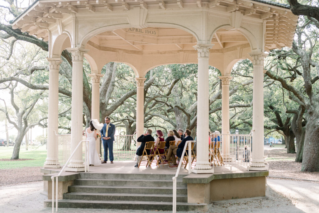 A beautiful micro wedding taking place under the gazebo in White Point Garden, Charleston. The bride looks stunning in a simple white dress, with her short veil gently blowing in the breeze. The groom is dapper in a blue suit, and the live oaks gracefully arch over the gazebo, creating a serene atmosphere for this intimate ceremony.