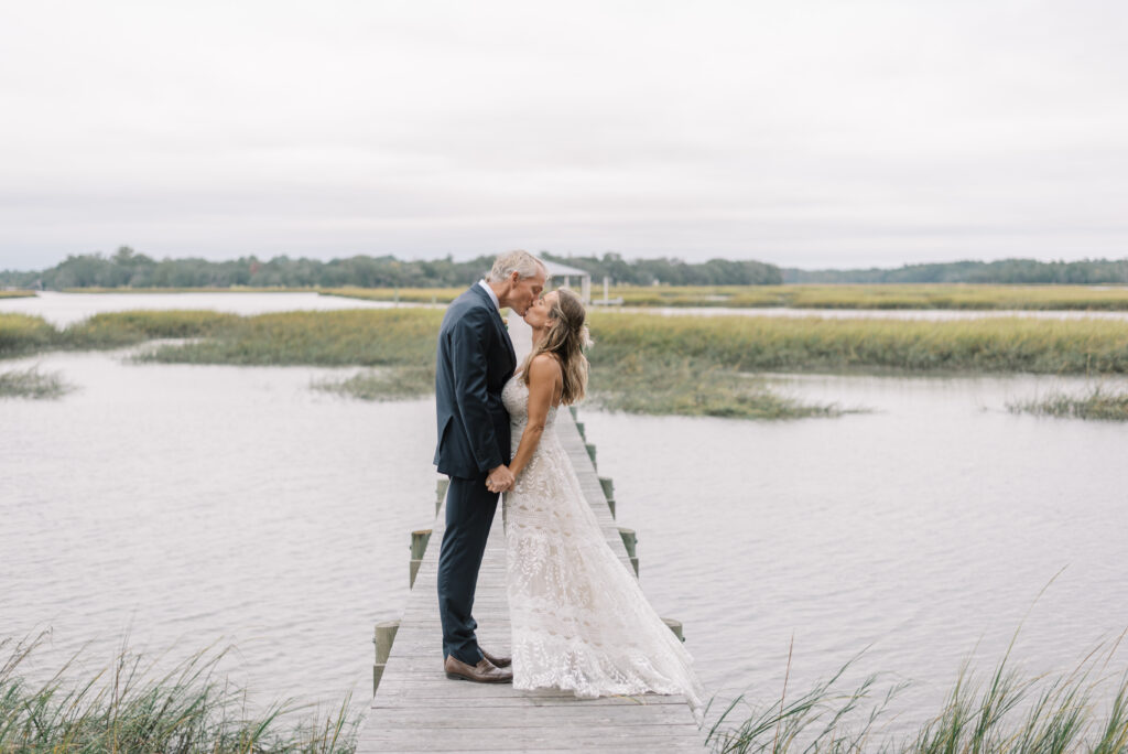 Captivating love: A heartwarming scene unfolds on a picturesque dock, where the groom, looking dashing in a navy suit, tenderly kisses his bride. She is a vision of beauty, adorned in a boho lace dress, her flowing locks half up, adorned with a delicate flower. Amidst the tranquil backdrop of a marsh, their love blossoms in this moment.