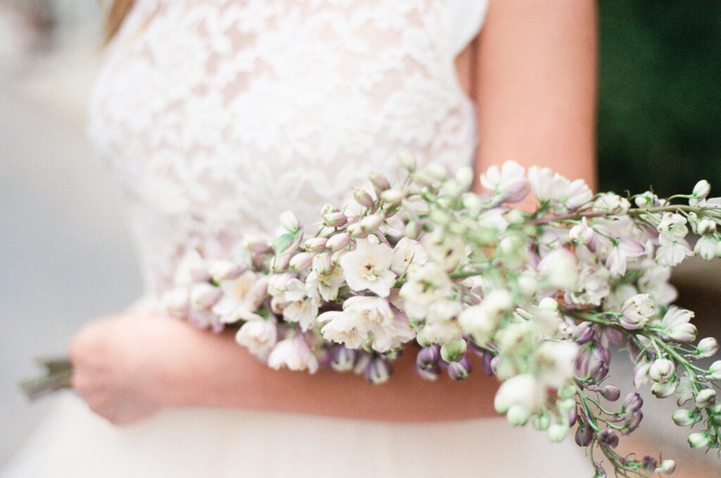 Breathtaking bride with bouquet: A captivating close-up image captured on film showcases the exquisite details of a bride's wedding day. The bride delicately holds her beautifully arranged bouquet, composed of white and lavender flowers, over her arm. The lace bodice of her dress peeks through, adding an element of enchantment to the scene. This timeless photograph captures the bride's grace, the artistry of her bouquet, and the intricate beauty of her attire, making it a captivating moment frozen in time.