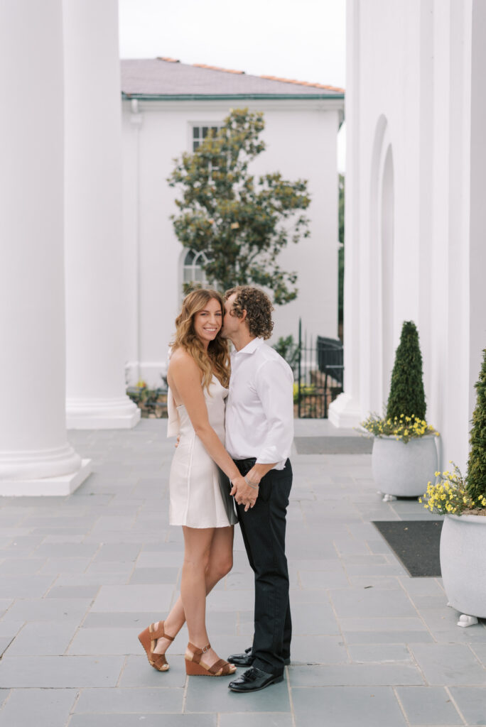 man whispers into his fiancé's ear as she smiles at the camera