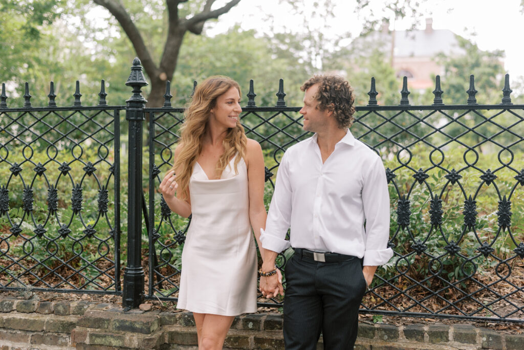 couple hold hands and smile at each other in front of a cast iron fence in Charleston