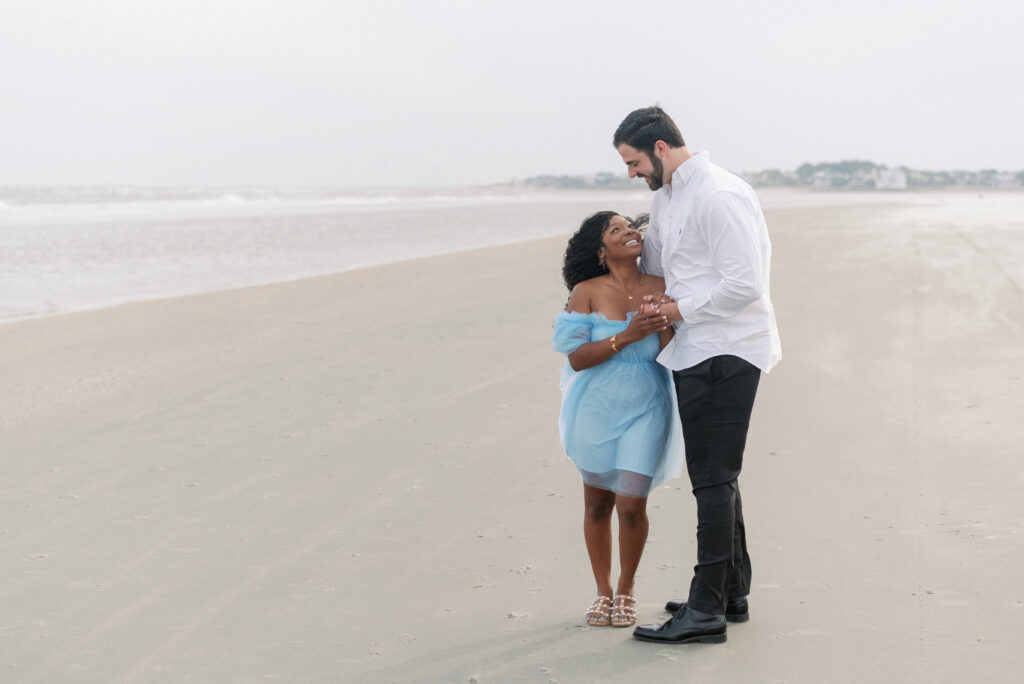 A heartwarming image of a biracial engaged couple sharing a tender embrace on the picturesque Isle of Palms beach. The groom-to-be looks dashing in black slacks and a crisp white shirt, exuding timeless elegance. The bride-to-be looks radiant in a captivating blue tulle dress, embodying grace and beauty. Their loving embrace captures the essence of their deep connection and anticipation for their upcoming union. This captivating moment, beautifully photographed, celebrates their love story and the joyous journey they are embarking upon together.