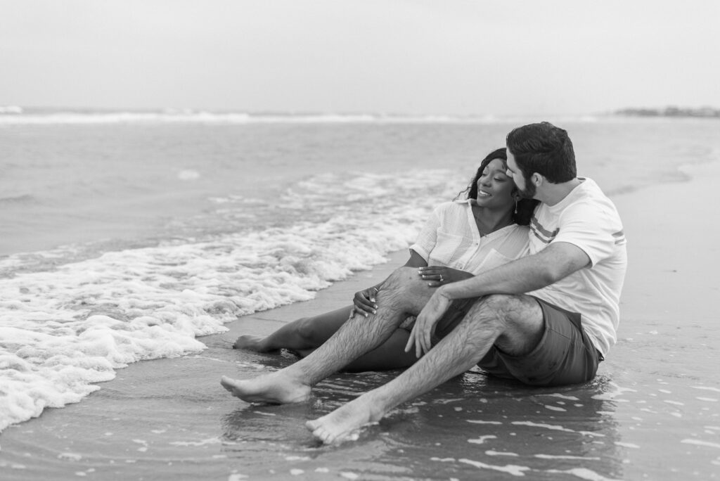 black and white portrait of an engaged couple sitting together on the beach in the water