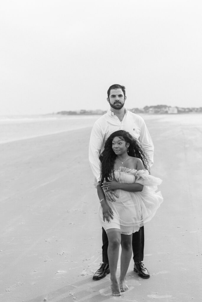 A captivating black and white image of an engaged couple standing together on the picturesque Isle of Palms beach. The bride-to-be leans back against her partner, gazing thoughtfully towards the glistening water, while the groom-to-be confidently meets the camera's gaze with a captivating expression. Their intimate connection and shared anticipation for their future are beautifully portrayed in this timeless snapshot. Expertly captured on film, this image showcases their love and the serene beauty of the beach.