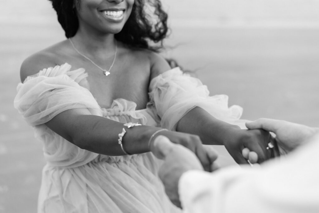 A captivating black and white close-up image of an engaged couple joyfully holding hands and spinning on the sandy beach. Their intertwined fingers and radiant smiles convey their deep love and happiness as they embrace the moment together. Expertly captured on film, this delightful snapshot encapsulates the excitement and romance of their engagement. The timeless black and white aesthetic adds a touch of elegance and nostalgia to this enchanting scene.