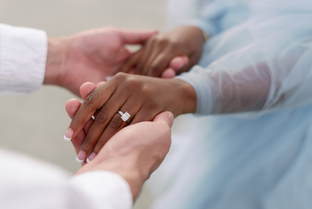 A captivating close-up image of an engaged couple holding hands, showcasing their love and commitment. The woman's hands are adorned with a flawless French manicure, beautifully highlighting her stunning emerald-cut engagement ring. She radiates elegance in a graceful long-sleeve light blue tulle dress, symbolizing purity and romance. This enchanting moment captured in the photograph is a testament to their joyous journey towards a lifetime of love and togetherness.