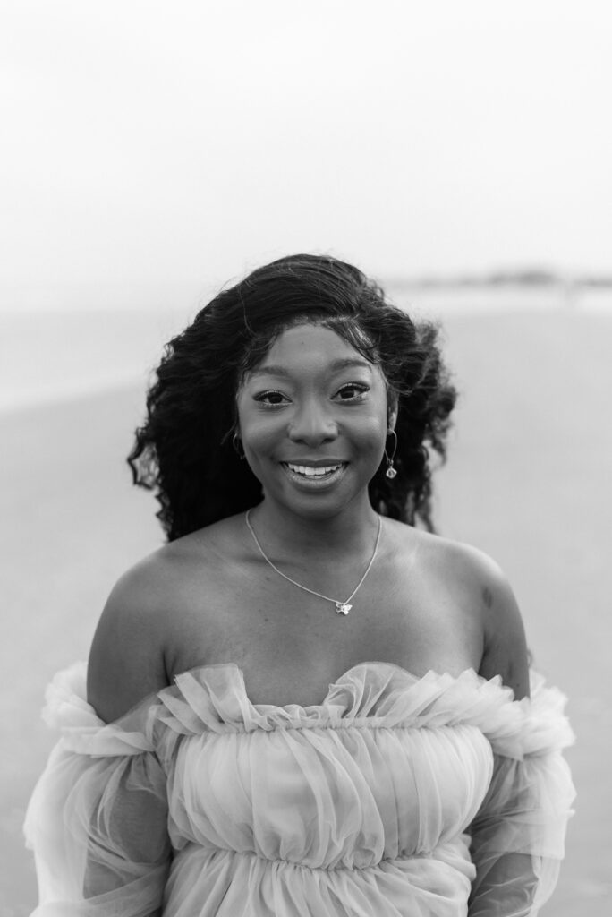 A captivating black and white photograph captured by The Wild Elopement, featuring a radiant woman adorned in a stunning tulle dress, standing gracefully on the beautiful Isle of Palms beach, as she beams with a genuine smile directed towards the camera.