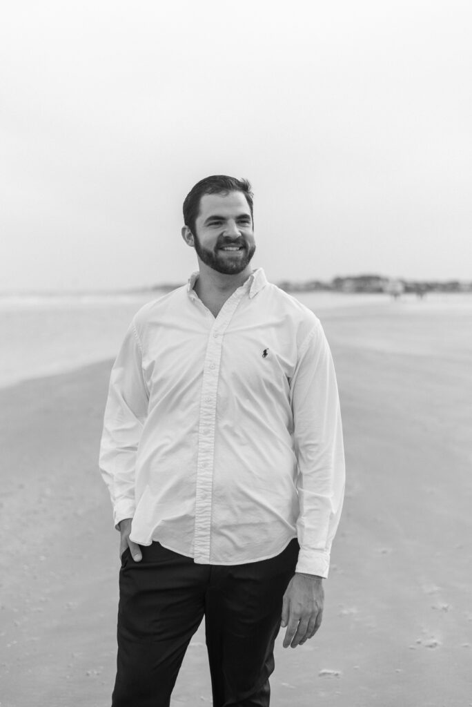 A captivating black and white photograph by The Wild Elopement, featuring a dashing man dressed in black slacks and a crisp white button-up shirt, standing on the picturesque Isle of Palms beach. With a radiant smile, he gazes away from the camera, exuding a sense of joy and tranquility.