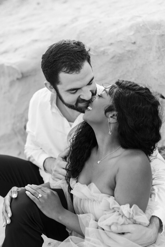 A captivating black and white close-up image of an engaged couple sitting in the soft sand, their faces inches apart, and their eyes locked with anticipation of a tender kiss. The intimate moment captured in this photograph radiates love, passion, and the promise of a lifelong commitment. The monochromatic tones lend a timeless and romantic feel to the scene, enhancing the emotional connection between the couple. This enchanting image beautifully encapsulates the depth of their affection and the excitement of their journey ahead.