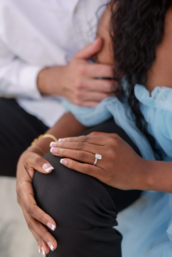 A captivating close-up image of an engaged couple seated side by side, their hands intertwined with the woman's delicate fingers resting on the man's knee, proudly displaying her exquisite diamond engagement ring. The intricate details of the ring sparkle in the light, symbolizing their love and commitment.