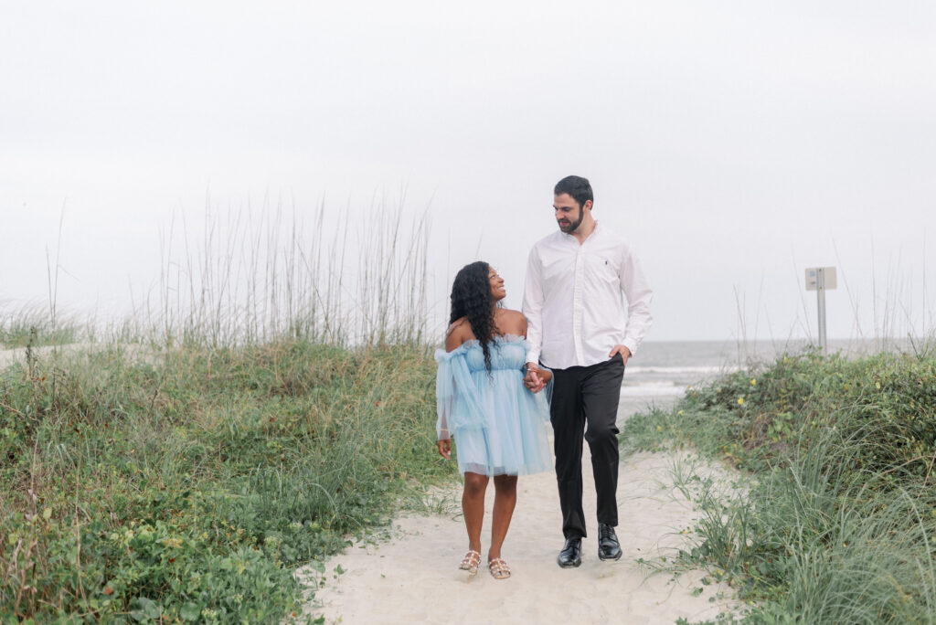 A delightful scene captured in this image as an engaged couple strolls hand in hand along a sandy path, moving away from the picturesque Isle of Palms beach. Their smiles radiate warmth and happiness, reflecting the deep connection they share. The path ahead symbolizes their journey together, filled with love, adventures, and cherished moments. This engaging photograph beautifully captures the essence of their relationship and the promise of a bright future.