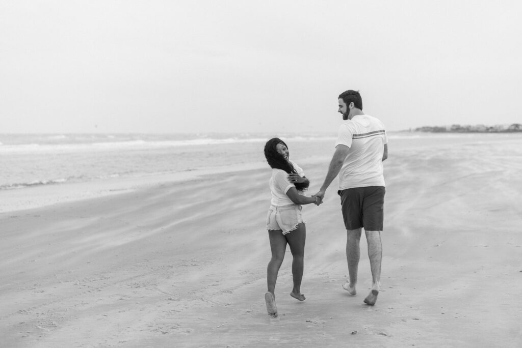 Engaged couple running on windy beach, captured in black and white.