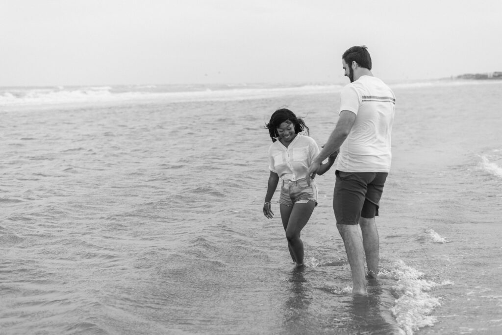 Engaged couple playing in the ocean together captured in black and white