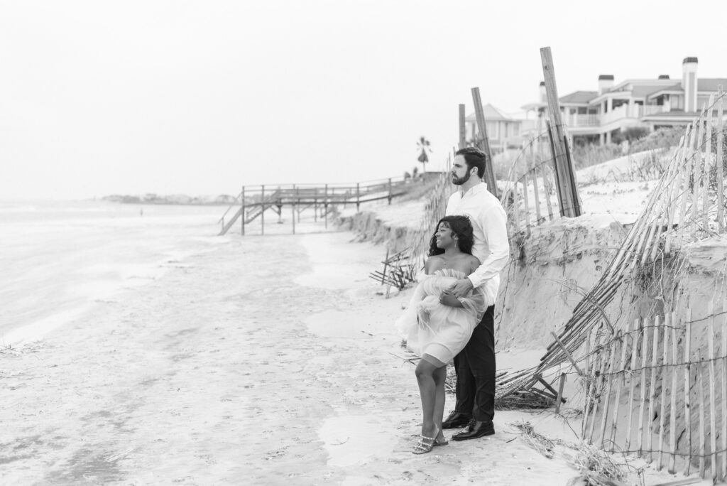 A timeless black and white photograph of a couple embracing on the picturesque Isle of Palms beach. The woman is adorned in a charming short tulle dress, while the man exudes elegance in black slacks and a crisp white shirt.