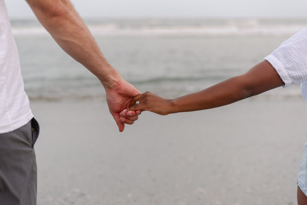 close up of an engaged couple holding hands on the beach looking towards the water