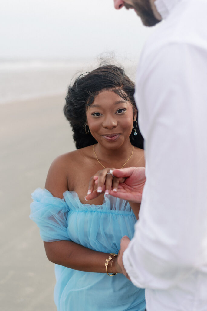 A captivating image of a biracial engaged couple standing together on the enchanting Isle of Palms beach. The groom-to-be exudes timeless charm in black slacks and a crisp white shirt, while the bride-to-be captivates with her radiant smile and direct eye contact with the camera. She looks stunning in a captivating blue tulle dress that complements the coastal backdrop. Their love and excitement for their journey together are beautifully showcased in this moment, skillfully captured by the photographer. This delightful snapshot celebrates their unique connection and anticipation for their upcoming union.