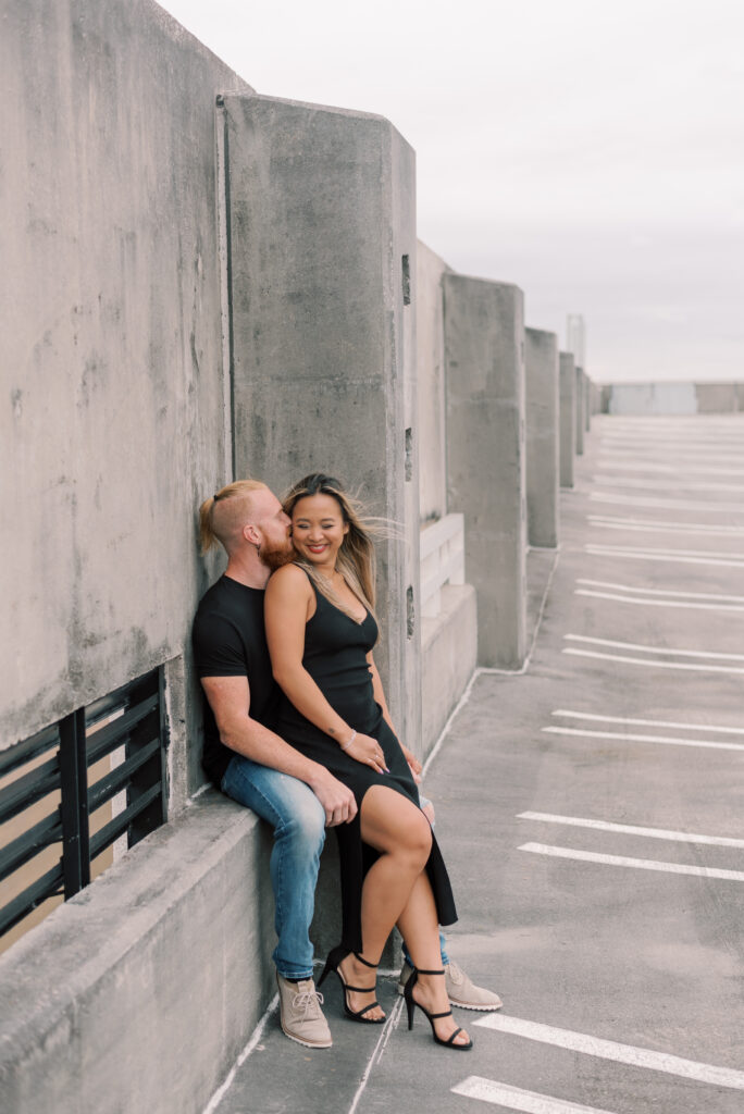 couple sitting on a parking garage ledge while the man whispers into the woman's ear