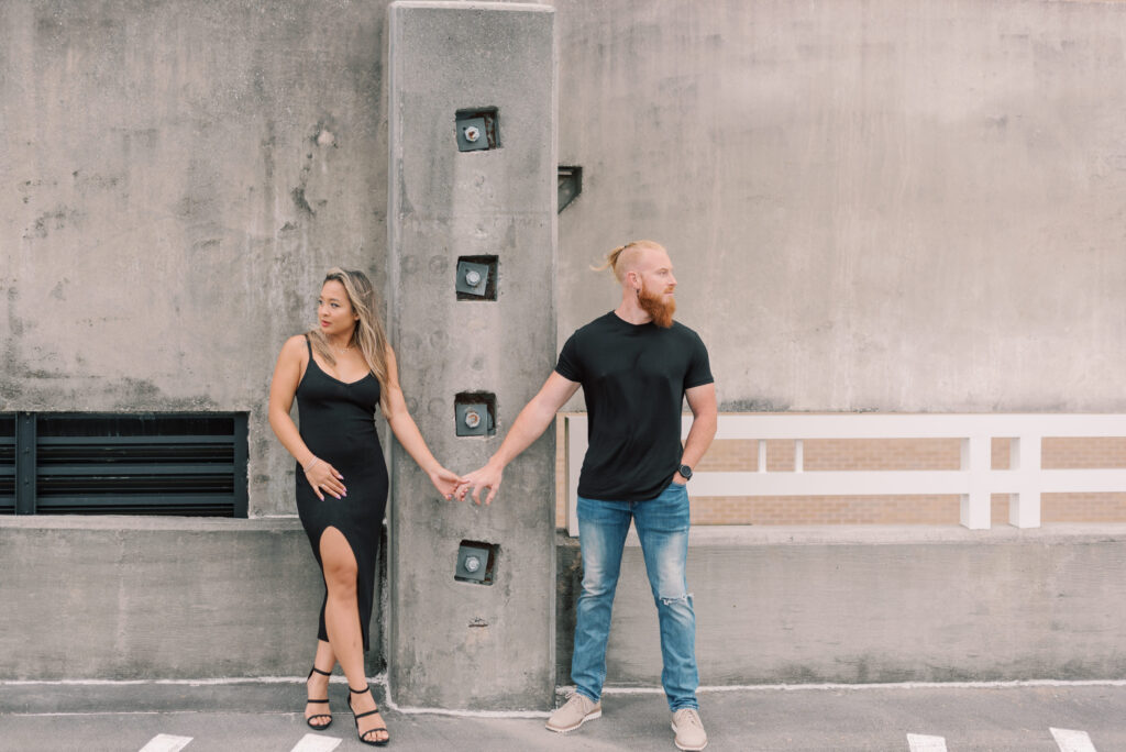 couple standing holding hands as they look away from each other with a concrete wall behind them