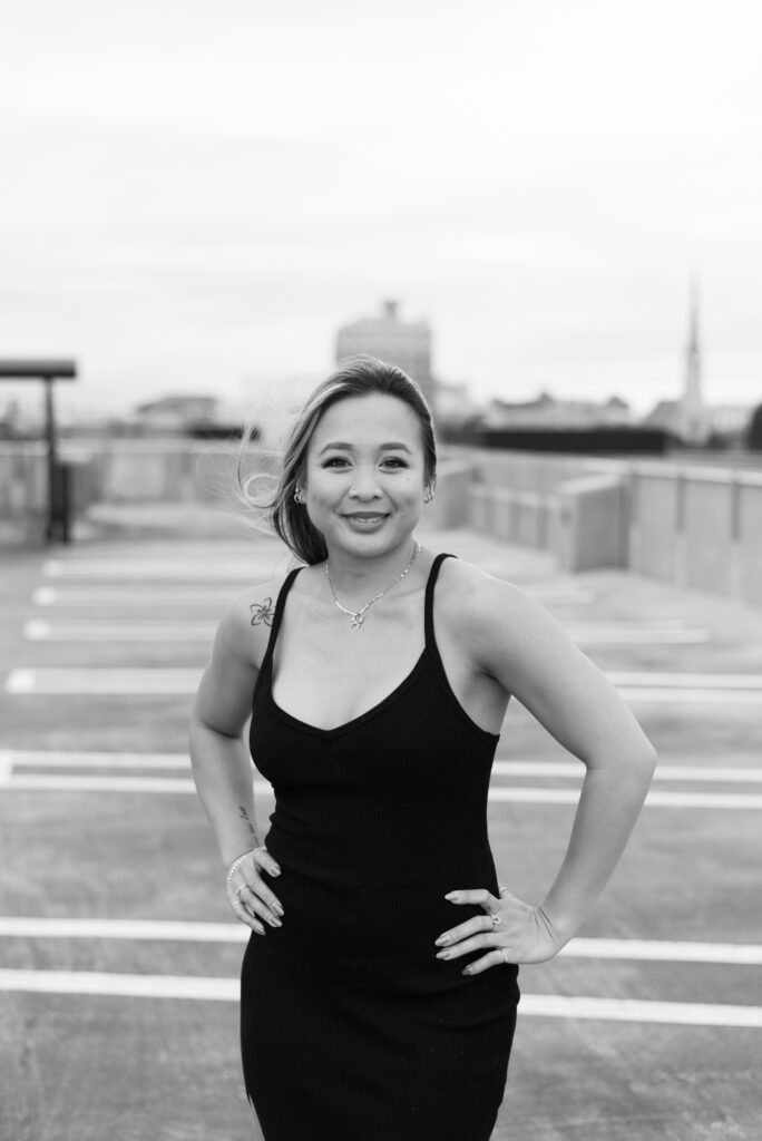 woman in a black dress, exuding elegance, standing on a parking garage against a black and white backdrop.