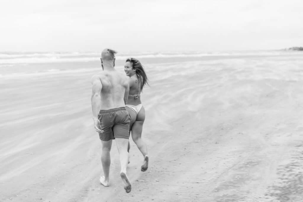 couple running across a windy beach while sand blows holding hands