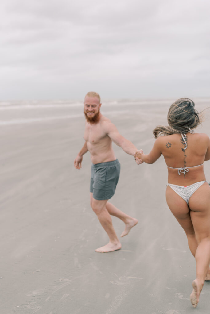 couple running and laughing at each other across a windy beach while sand blows