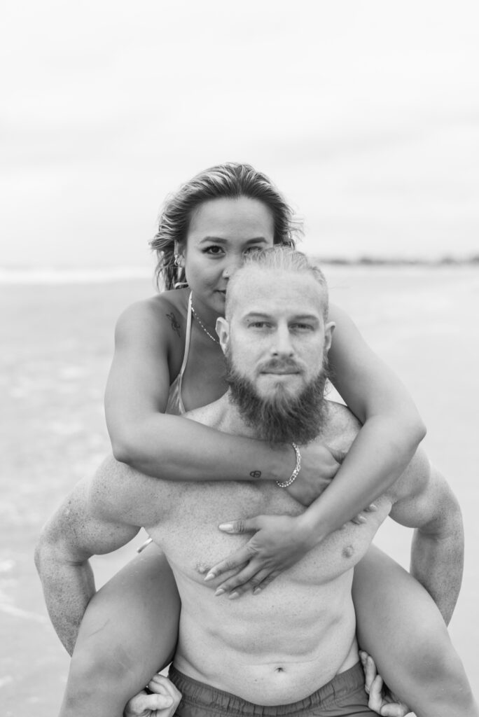 black and white portrait of a man and woman staring into the camera while he holds her piggyback