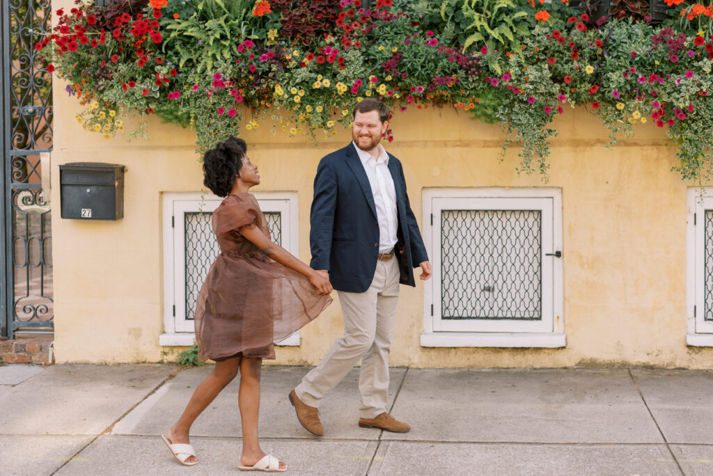 Couple hold hands walking in front of  a yellow house in historic Charleston