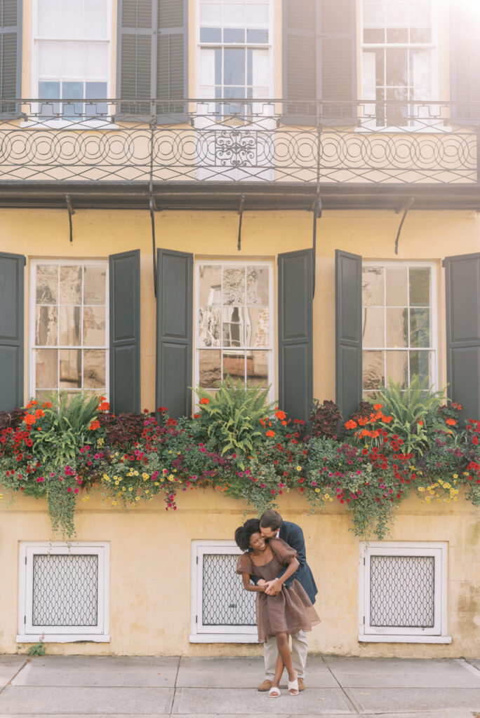 Man kisses his fiancé on the cheek in front of a yellow house in historic Charleston