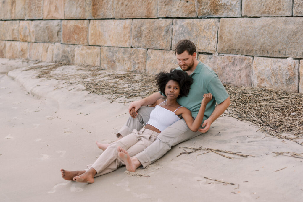 woman lays between her fiancé's legs sitting on the beach