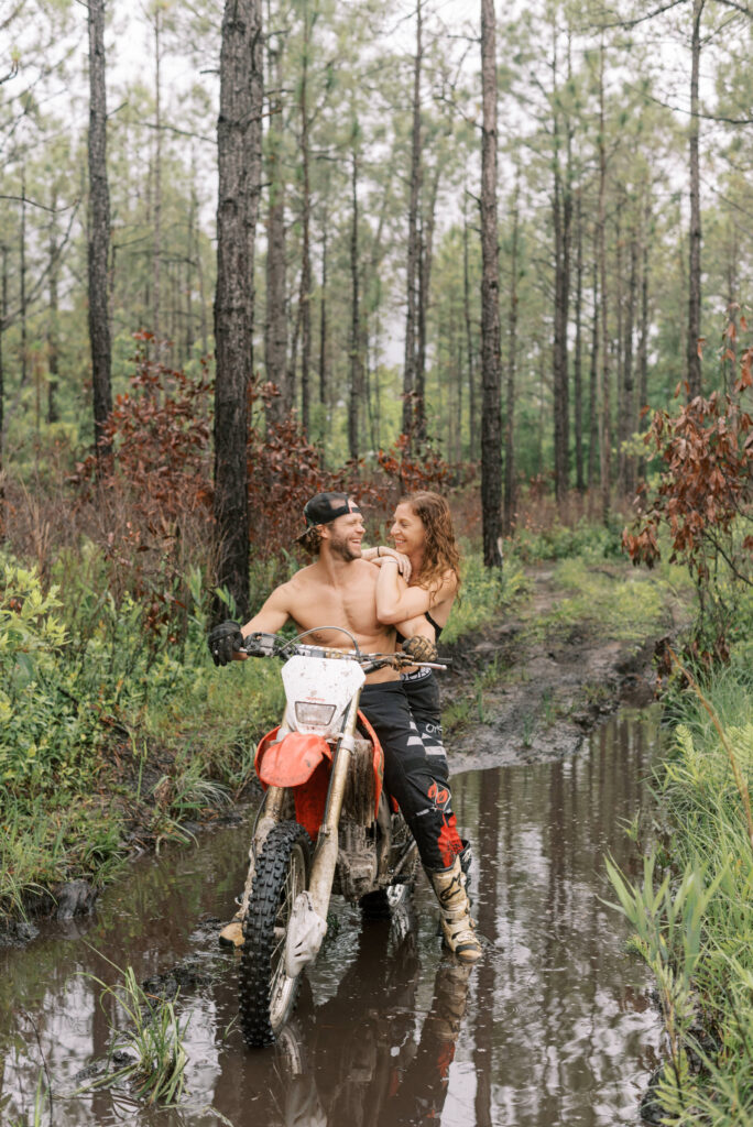 Couple sits together on a dirt bike in the middle of a mud hole in the woods