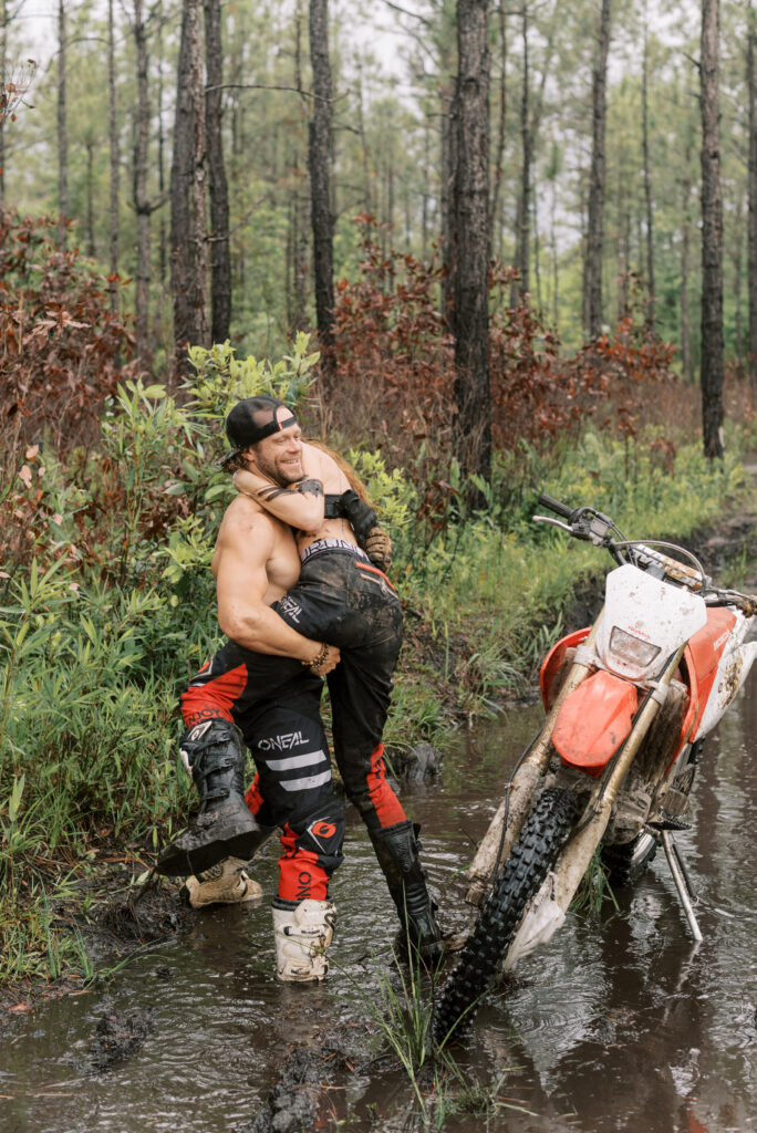 man picks his girlfriend up in the middle of a mud pit in the woods beside a dirt bike