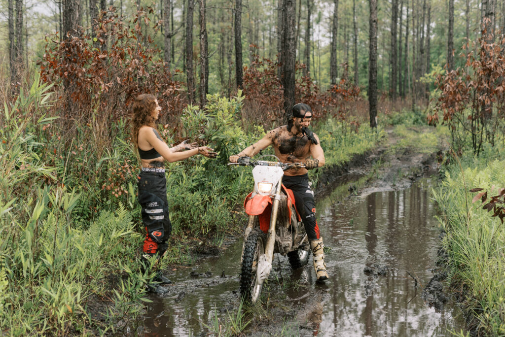 woman throws a clump of mud at her boyfriend while he rides his dirt bike through a mud hole