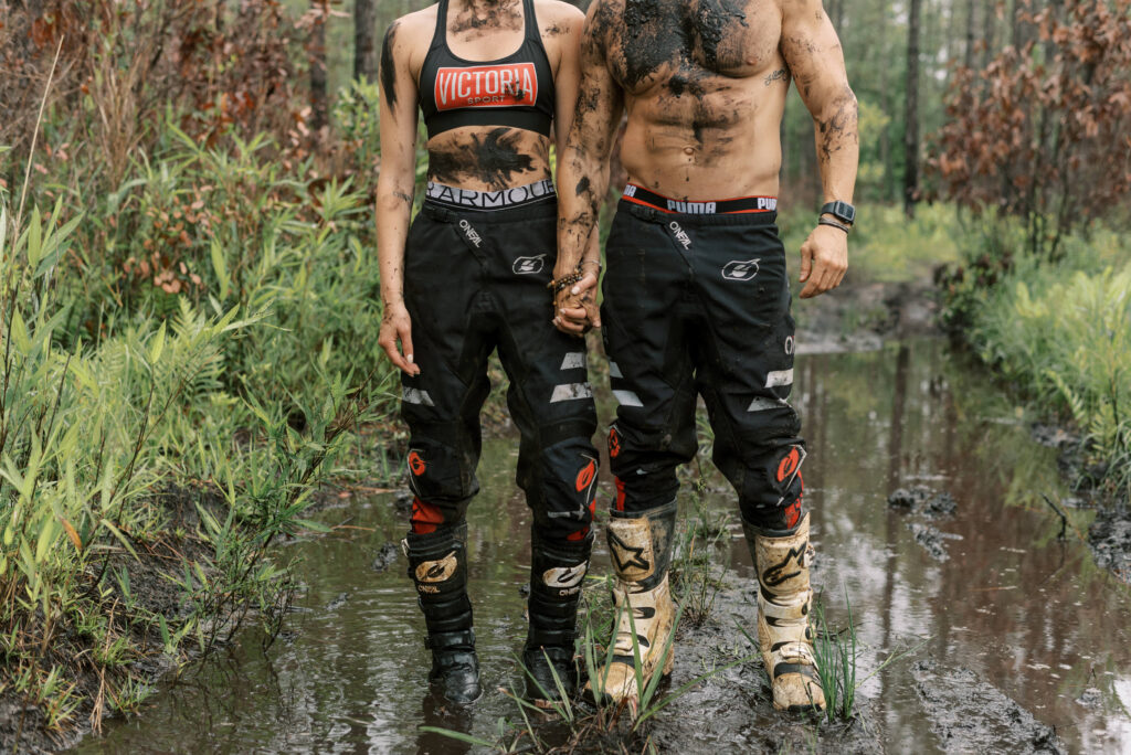 Couple covered in mud stand in a mud hole in just their oneil motorcycle pants and puma and under armour clothes